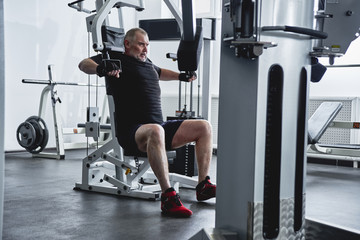 Fototapeta na wymiar Adult gray-haired man trains on fitness equipment in the gym, pumps legs and arms muscles, loses weight. Concept of healthy lifestyle in old age