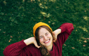 Above view image of a happy blonde young woman smiling broadly with a healthy toothy smile, wearing a red sweater, and yellow hat, posing on greenery nature background in the park.