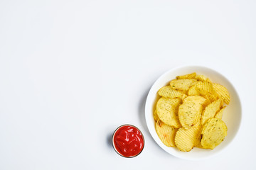 Potato chips in a white bowl next to tomato ketchup on a white background. Top view.