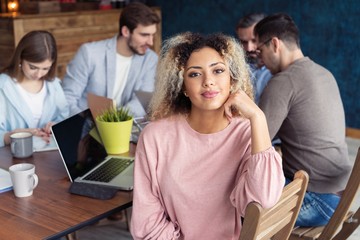Happy to be in team. Beautiful cheerful African woman looking at camera with smile while sitting at the office table with her coworkers.