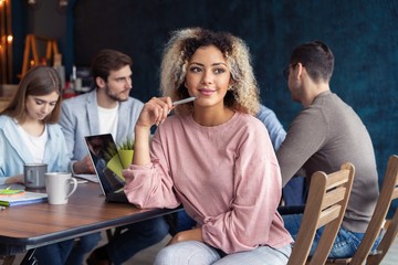 Happy to be in team. Beautiful cheerful African woman looking at camera with smile while sitting at the office table with her coworkers.