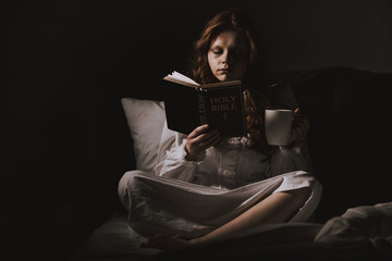 demonic woman in nightgown holding cup and reading holy bible on bed