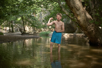 Fototapeta na wymiar Smiling teenage boy posing like bodybuilder in shallow water in forest in mediterranean country in summer time, sunny relaxed mood fun smile happiness