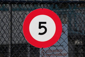 View of round white road sign with red border indicating speed limit of 5 km/h, on industrial metal mesh fence