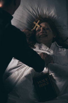 exorcist with bible and cross standing over demoniacal girl in bed