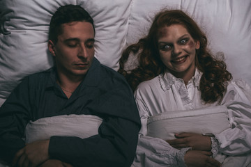 top view of creepy smiling female demon lying in bed with sleeping man