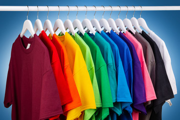 row of many fresh new fabric cotton t-shirts in colorful rainbow colors hangng on clothes rail in...