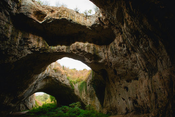 High arches of a huge stone cave with round holes at the top, a tourist road with a fence inside...