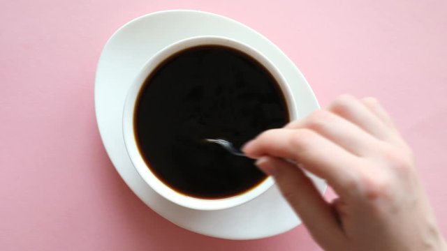 Woman's hand stir sugar in cup of coffee on pink background