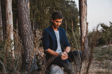 Handsome man posing outdoors in forest sits on a log, wearing checked jacket. Place for text or advertising