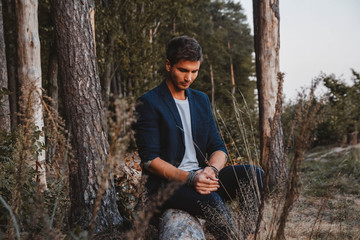 Fototapeta premium Handsome man posing outdoors in forest sits on a log, wearing checked jacket. Place for text or advertising