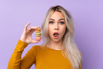 Teenager girl over isolated purple background holding colorful French macarons with surprise expression
