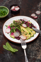 selective focus of fresh radish salad with greens and avocado on plate served on weathered surface with cutlery and ingredients