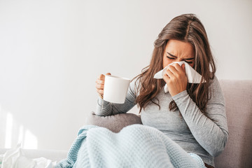 Sick woman covered with a blanket lying in bed with high fever and a flu, blowing her nose. Pills and glass of water on the table. Woman sneezing in a tissue in the living room - obrazy, fototapety, plakaty