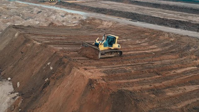 Top drone view on crawler excavator controlled by operator. Bulldozer digging sand in sandy quarry on construction apartment buildings sleeping city area. Preparation of site for apartment building.