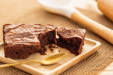 Brownies cake in a wooden plate with a golden spoon, large cooking spoon and rolling pin in the background on natural table mat..