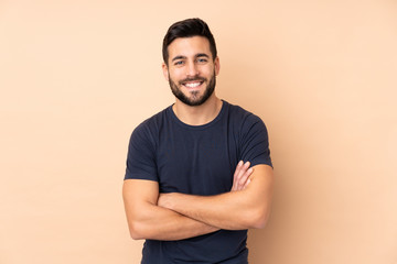Caucasian handsome man isolated on beige background keeping the arms crossed in frontal position - obrazy, fototapety, plakaty