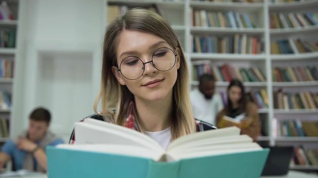 Pretty calm concentrated young woman in glasses reading book in the university library