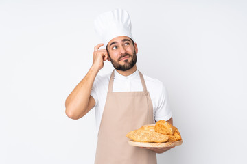 Male baker holding a table with several breads isolated on white background having doubts and with confuse face expression