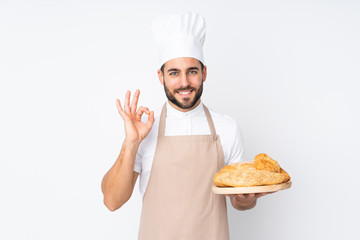 Male baker holding a table with several breads isolated on white background showing an ok sign with fingers