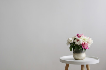 Close up shot of beautiful decorative glass vase with bouquet of peonies on white coffee table on foreground and blank wall with a lot of copy space for text on background.