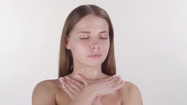 Portrait shot of beautiful young woman standing isolated on white background and massaging lotion into her neck and collarbone while looking at camera