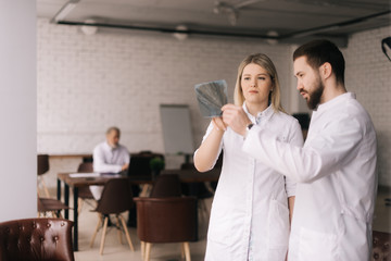 Two young dentists are examining the patient's jaw X-ray in modern meeting office room of the hospital.. There's an adult experienced doctor sitting in the background.