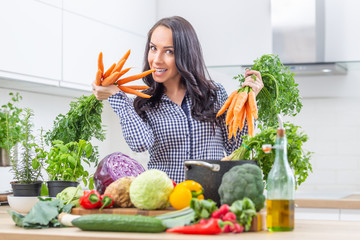Playful young woman in her kitchen holding fresh carrot in both hands - diet vegetable and heath concept