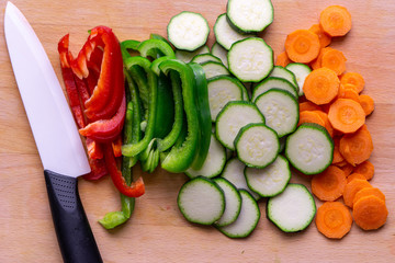 Cut vegetables on a wooden cutting board: carrots, zucchini, red and green paprika. There is a knife near the vegetables. 