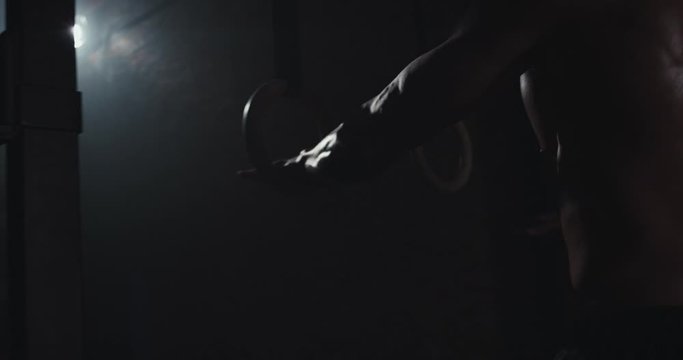 In a dark gym class athletic strong guy starting his hard work training , he balancing on the sport rings. 4k
