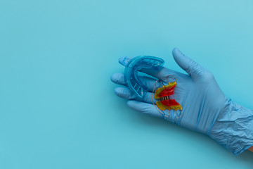 Hand of medic wearing blue latex glove holding myofunctional trainer and retainer orthodontic appliance on blue background. Teeth correction concept