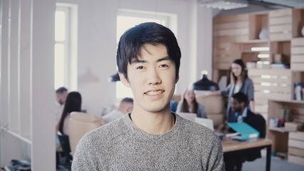 Obraz premium Portrait of young successful Asian businessman smiling at busy office. Handsome male manager looking at camera posing 4K