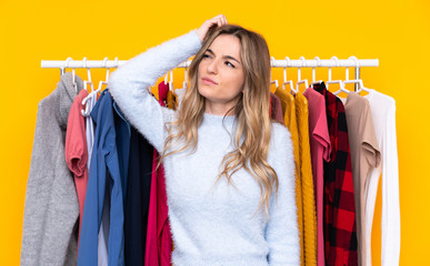Young woman in a clothing store over isolated yellow background having doubts and with confuse face...