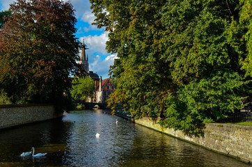 Naklejka premium Bruges, Belgium. August 2019. View towards the historic center from the bridge with the Sashuis lock. The canal leads the eye towards the city. Large green foliage on the sides.