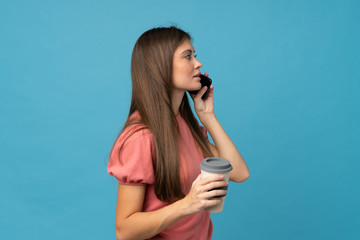 Young woman over isolated blue background holding coffee to take away and a mobile