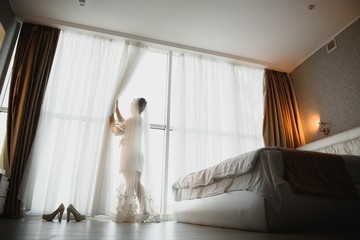 Fototapeta na wymiar Portrait of beautiful bride in white silk dressing gown with curly hairstyle and veil standing near window in bedroom, copy space. Bride's morning preparation. Back view