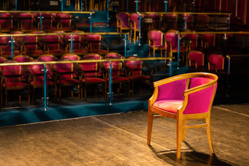 Red upholstered chair on a theater stage with reflectors ray with a empty blurred rows of chairs of theater hall , high contrast image. The show must begin.