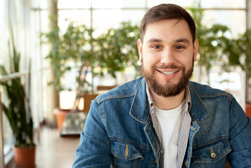 Obraz premium Portrait smiling handsome young man with beard in modern corporate environment near window. Smart man in office in casual wear, jean jacket. Attractive businessman freelancer smiling in cafe.