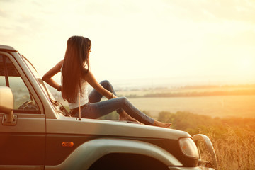 Attractive yong woman is sitting on the car's hood and looking at sunset. Rural evening background. - obrazy, fototapety, plakaty