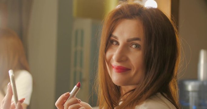 Attractive brunette young woman putting on dark red lipstick on her lips, looking in the mirror. Reflection of a lovely lady applying her makeup. The girl paints her lips with red lipstick and looks
