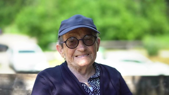 Portrait of an elderly sporty woman  with a blue cap on and rounded black glasses looking at camera and happily smiling. Slow motion of nice retired lady in the park. Cine lens.