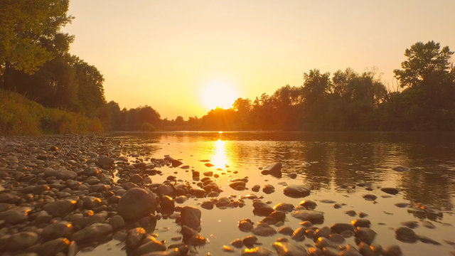 AERIAL, CLOSE UP: Rocky riverbank and water surface reflecting golden sky