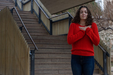 Portrait of caucasian young woman with closed eyes, keeps hands on chest, in the park, orange sweater and jeans, long curly hair. Place for your text in copy space.