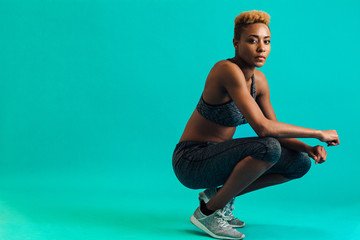 Portrait of a serious female athlete in sports bra and gym tights, squatting and looking at camera,...