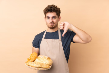 Male baker holding a table with several breads isolated on beige background showing thumb down sign