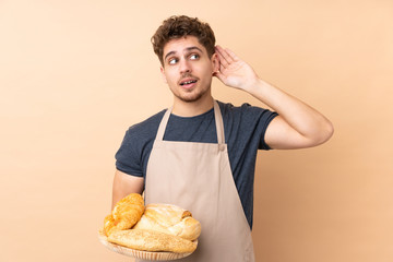 Male baker holding a table with several breads isolated on beige background listening something