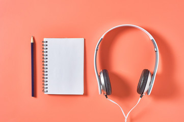 Modern white headphones with cable near blank paper notebook and pencil on orange background. Music concept. Copy space. Top view