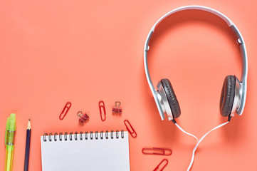 Modern white headphones with cable near blank paper notebook and other stationery things on orange background. Business or school concept. Copy space. Top view