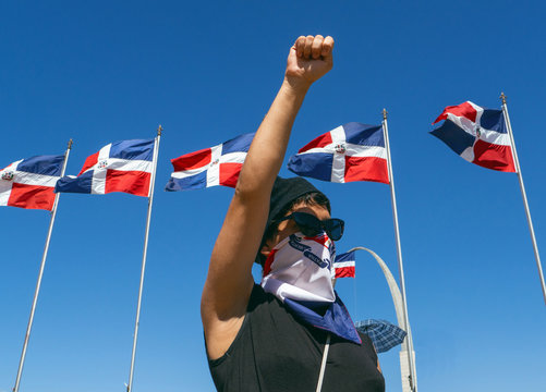 A girl with glasses, hat and scarf covering her mouth, fights for her rights with the Dominican flags and the blue sky in the background