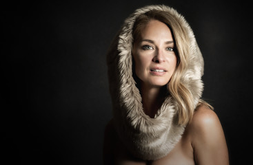 Glamour photo of a beautiful (middle aged) 45 years old blonde woman. With black background and room for text. She has bare shoulders and dressed in fur. 
